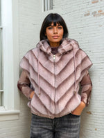 Woman in a blush pink sleeveless faux fur hoodie with chevron stripes and gold zip, layered over a sheer printed top and dark jeans.