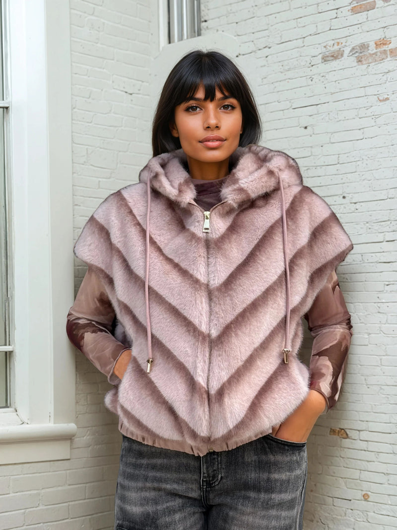 Woman in a blush pink sleeveless faux fur hoodie with chevron stripes and gold zip, layered over a sheer printed top and dark jeans.