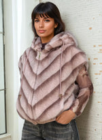 Model wearing a light mauve-pink faux fur hooded vest with drawstrings, paired with sheer floral long sleeves and faded denim, standing near a white wall.