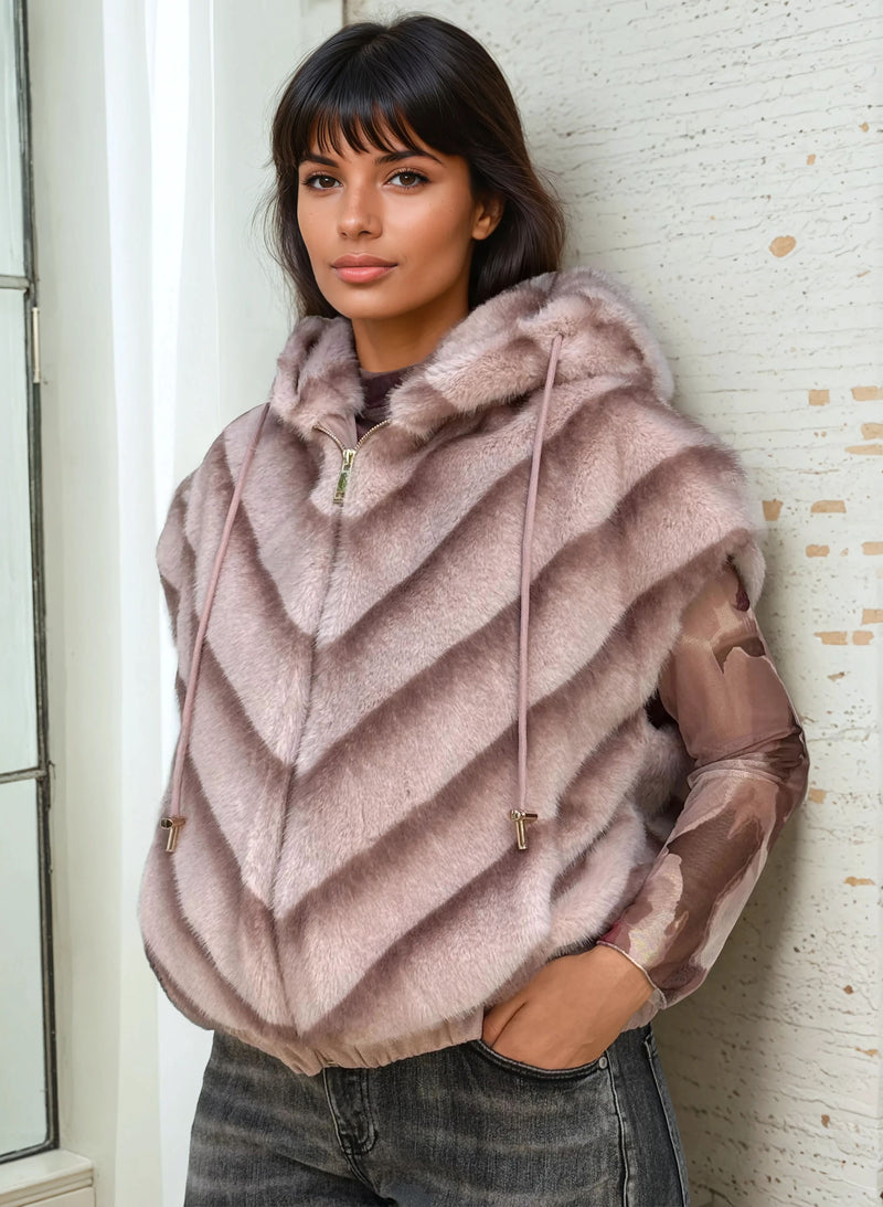 Model wearing a light mauve-pink faux fur hooded vest with drawstrings, paired with sheer floral long sleeves and faded denim, standing near a white wall.
