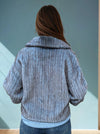 Back view of a blue ribbed faux fur jacket showing textured vertical lines and structured collar, worn with denim jeans.