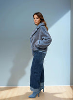 Full body view of a model in a blue ribbed faux fur short coat, styled with wide-leg cuffed jeans and pointed blue boots in natural light.