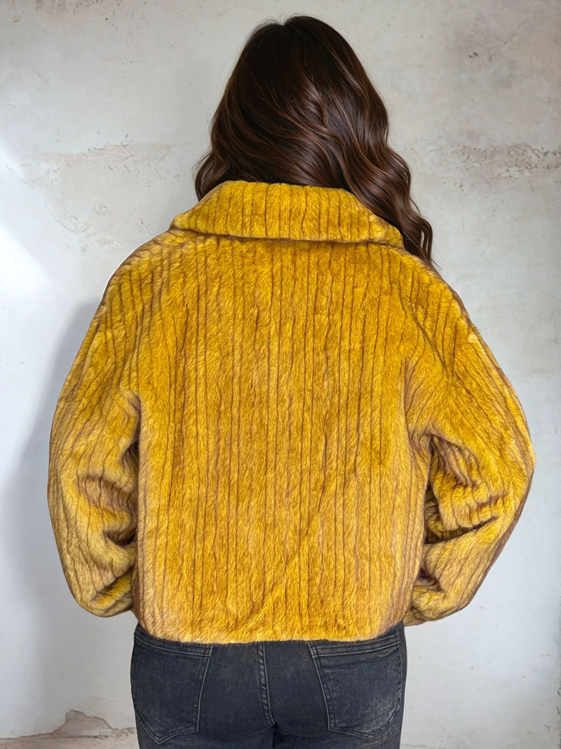 Back view of mustard yellow faux fur jacket highlighting ribbed texture and wide collar, modeled indoors with natural lighting
