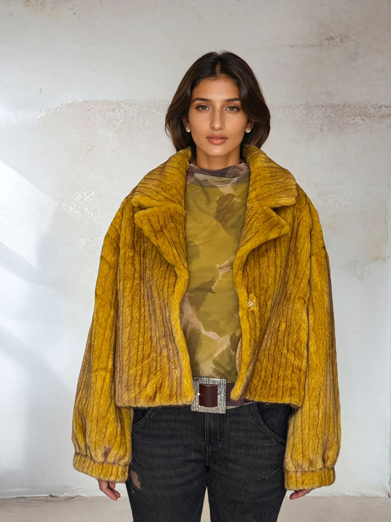 Woman wearing mustard yellow faux fur jacket with wide lapels and textured design, styled over a camouflage top and black jeans, standing against a neutral wall background.