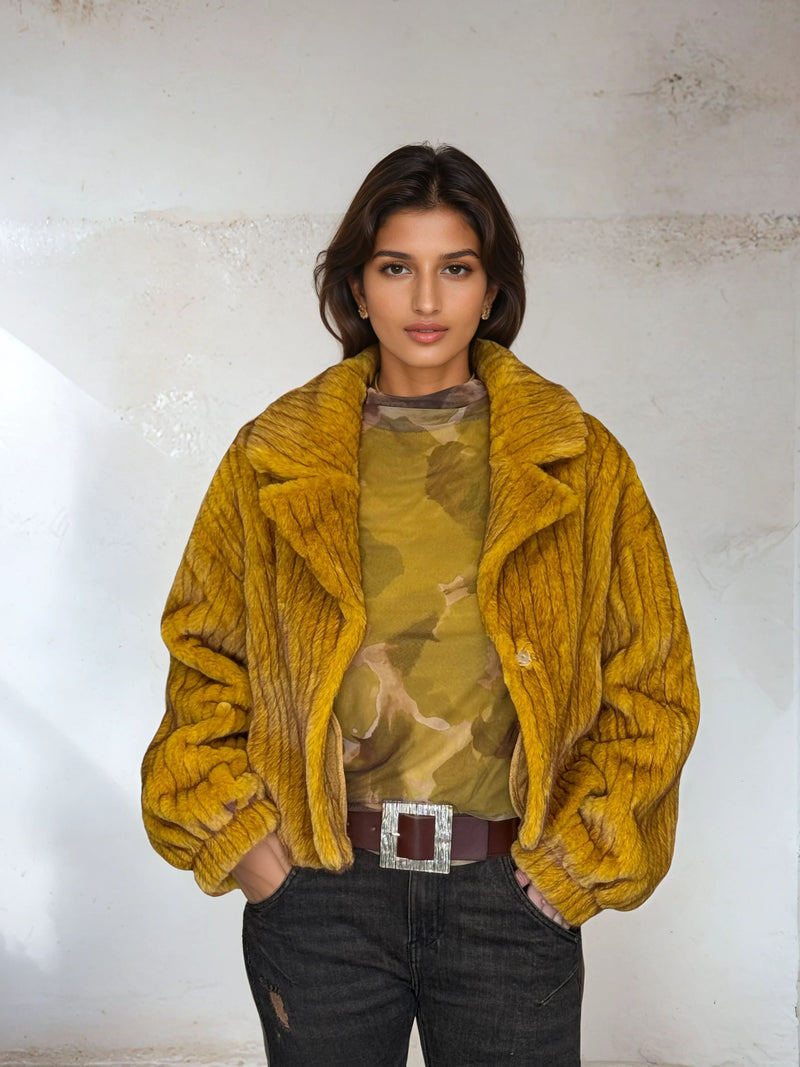 Model in mustard yellow plush fur coat with textured ribbed pattern, paired with camo top and dark denim jeans for a modern winter look.