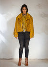 Full body shot of model in mustard yellow fur jacket, camouflage print top, black skinny jeans, and brown leather boots in a minimalist setting