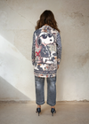 Full-length back view of a woman wearing a casual printed coat with Snoopy graphic and animal print design, paired with rolled gray jeans and black heels, posing in a minimal room.